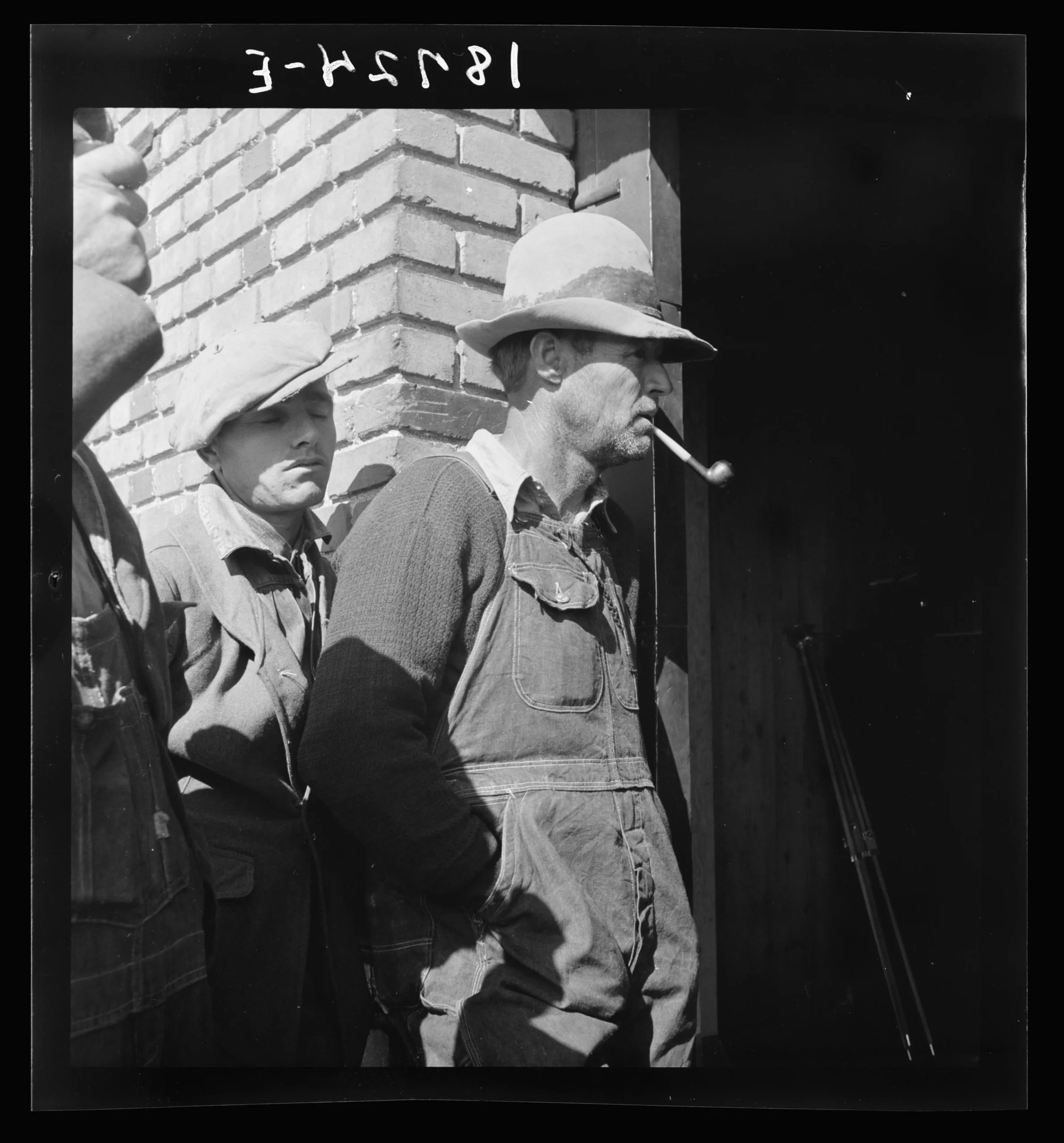 Photograph of three men standing outside of a darkened doorway on a brick building. The primary subject of the photo has a pipe in his mouth, a rumpled hat on his head, and is wearing overalls over a sweater and collared shirt combo.